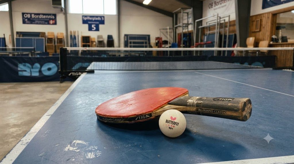 Bordtennisbat med rød belægning og bold på blåt bord i dansk klubhal