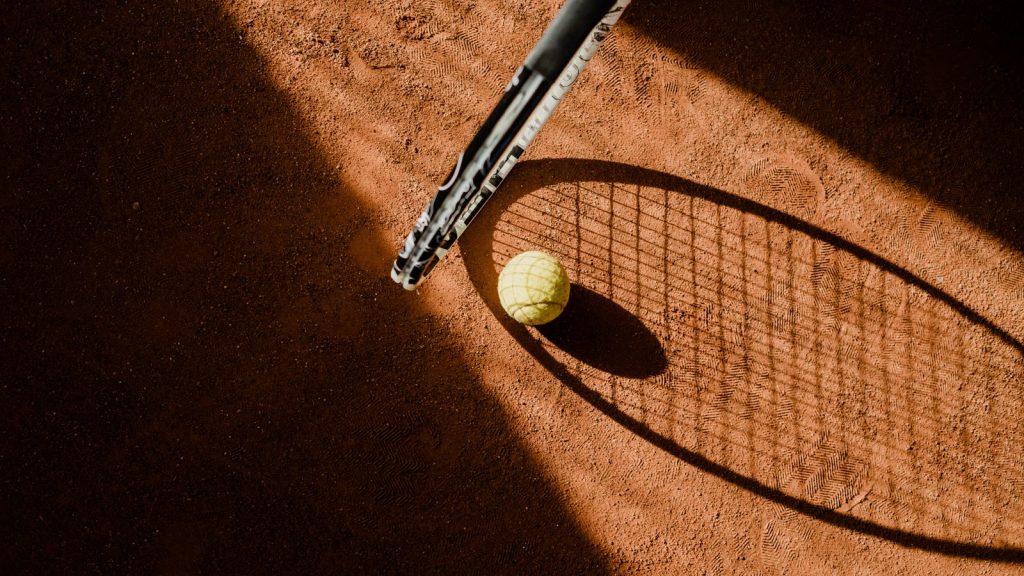 Tennisbold og skyggen af en ketsjer på en rød grusbane i lavt sollys
