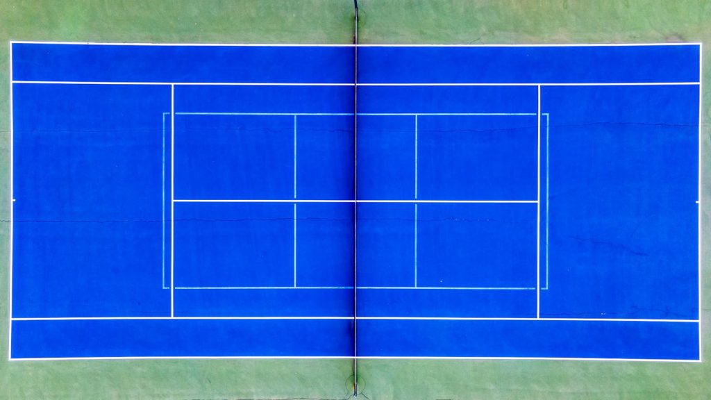 Blå hardcourt-tennisbane set lige oppefra med hvide banelinjer og nettet ned gennem midten