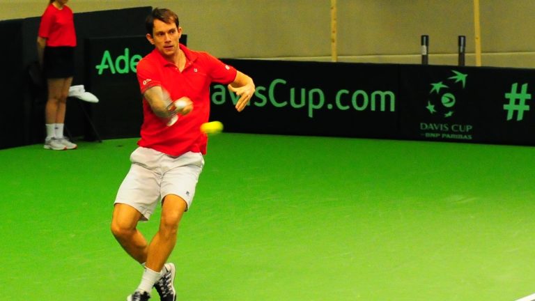 Frederik Løchte Nielsen slår forhånd i rød dansk landsholdstrøje foran DavisCup.com-banneret ved en indendørs Davis Cup-kamp