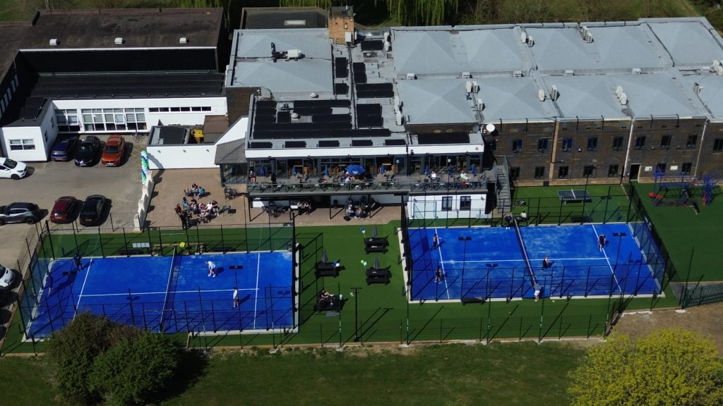 Luftfoto af to udendørs padelbaner med blåt underlag ved et klubhus med terrasse