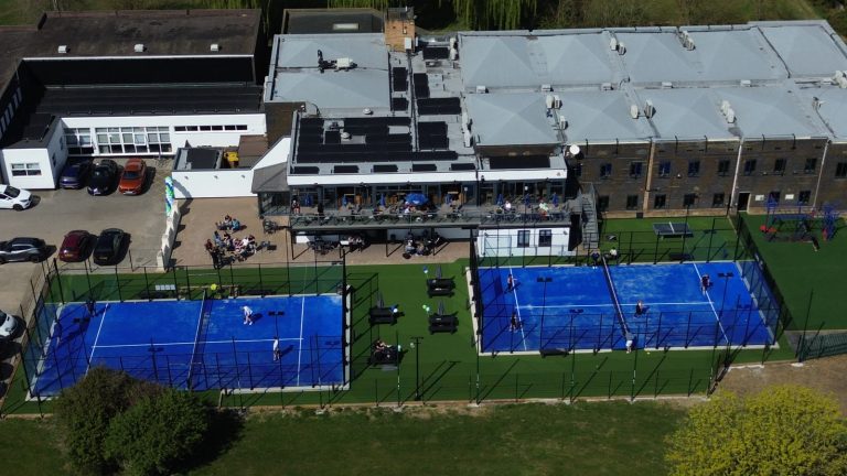 Luftfoto af to udendørs padelbaner med blåt underlag ved et klubhus med terrasse