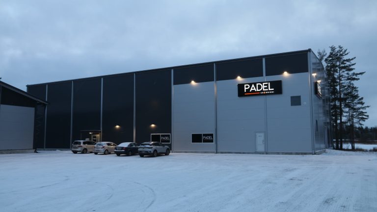 Facaden på en indendørs padelhal med oplyst PADEL-skilt og parkerede biler på en snedækket plads i skumringen