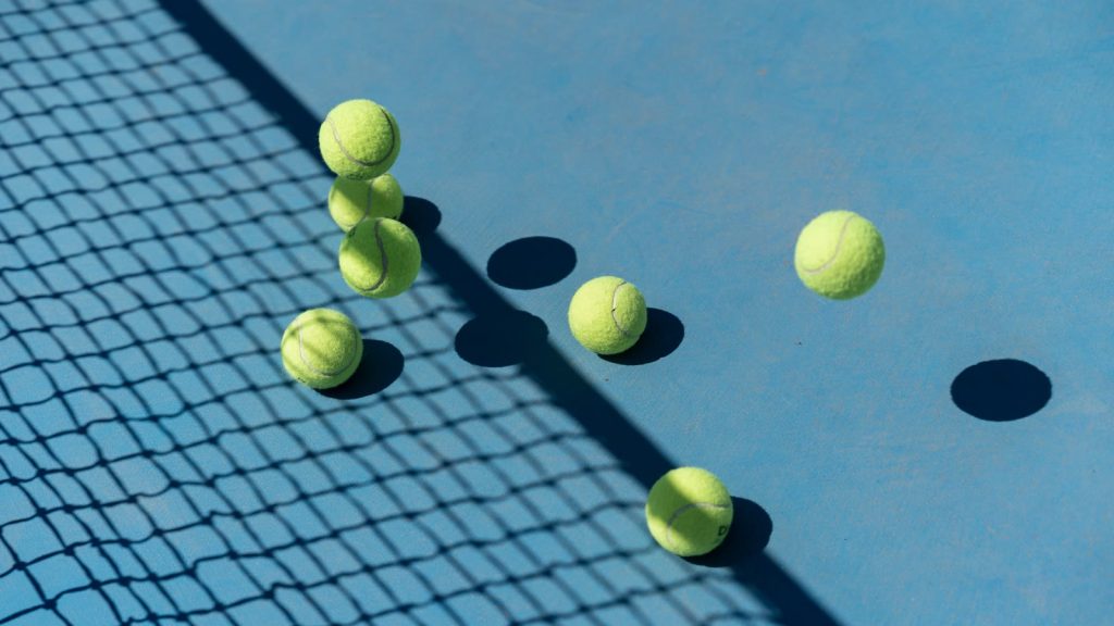 Tennisbolde, der hopper på en blå hardcourt-bane i skarpt sollys med nettets skygge hen over underlaget