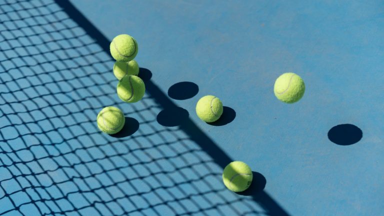 Tennisbolde, der hopper på en blå hardcourt-bane i skarpt sollys med nettets skygge hen over underlaget