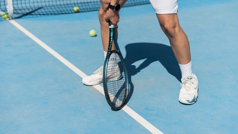 Tennisspiller med ketcher i hånden, set fra hofterne og ned, i klarposition ved baglinjen på en blå hardcourt-bane